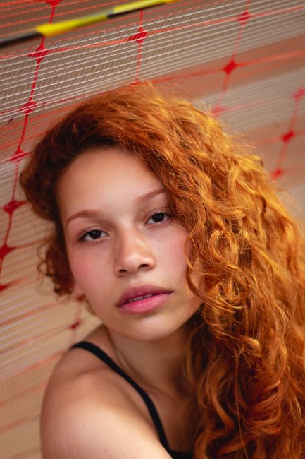 Close-up portrait of a young woman with vibrant red curly hair looking confidently at the camera.
