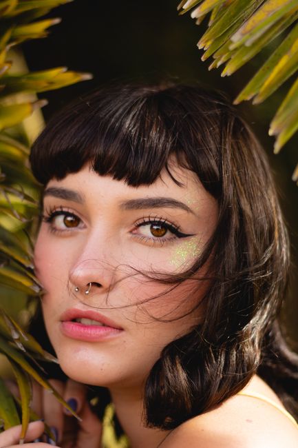 Stunning portrait of a young woman with brown eyes and septum piercing surrounded by tropical leaves.