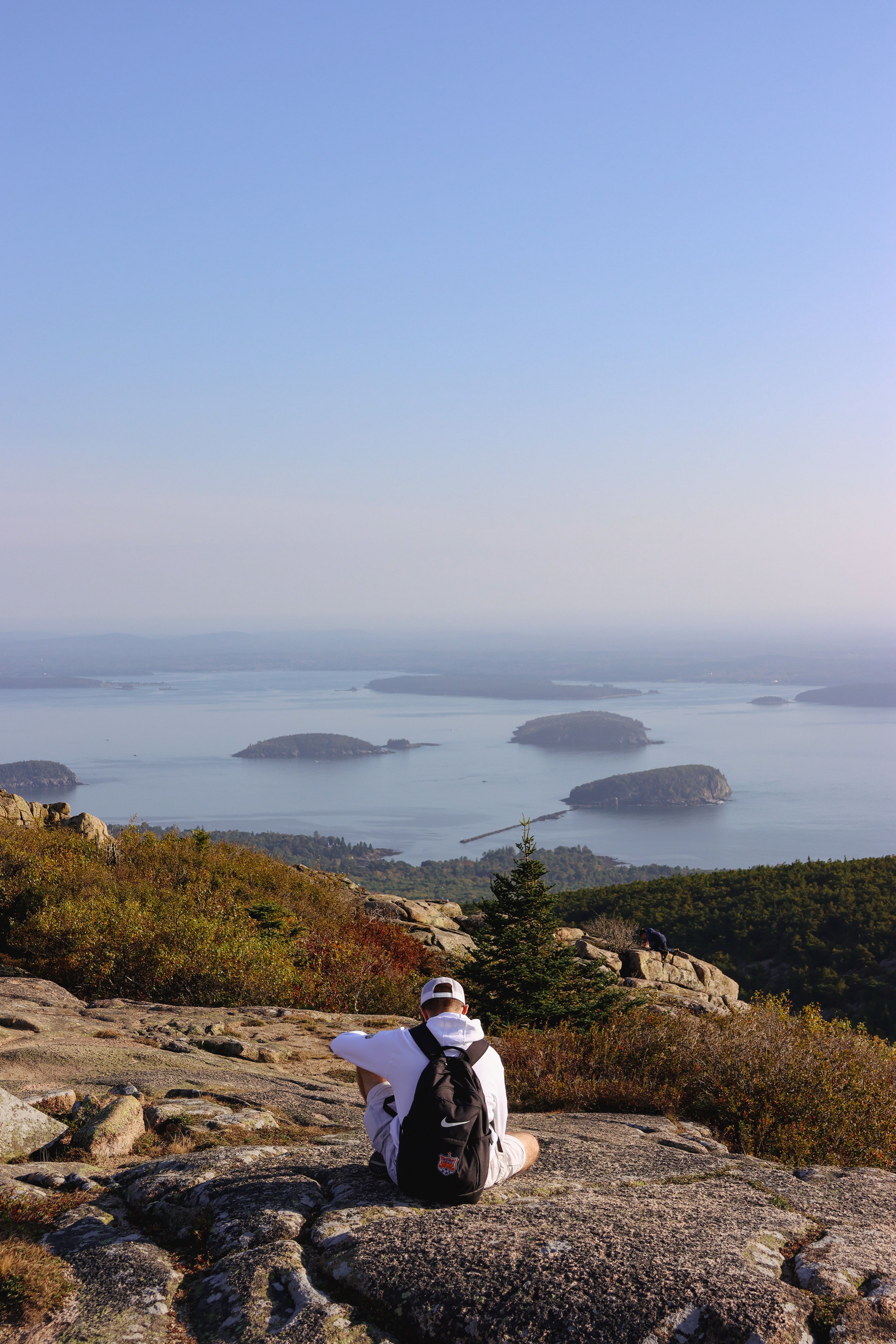 A person in a backpack sits on a hill overlooking Frenchman Bay, capturing the serenity of Maine's natural beauty.