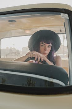 Stylish woman in a dark hat gazing through a vintage car's windshield on a sunny day.