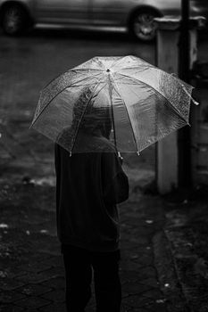 A person stands on a city sidewalk with an umbrella on a rainy day, captured in black and white.