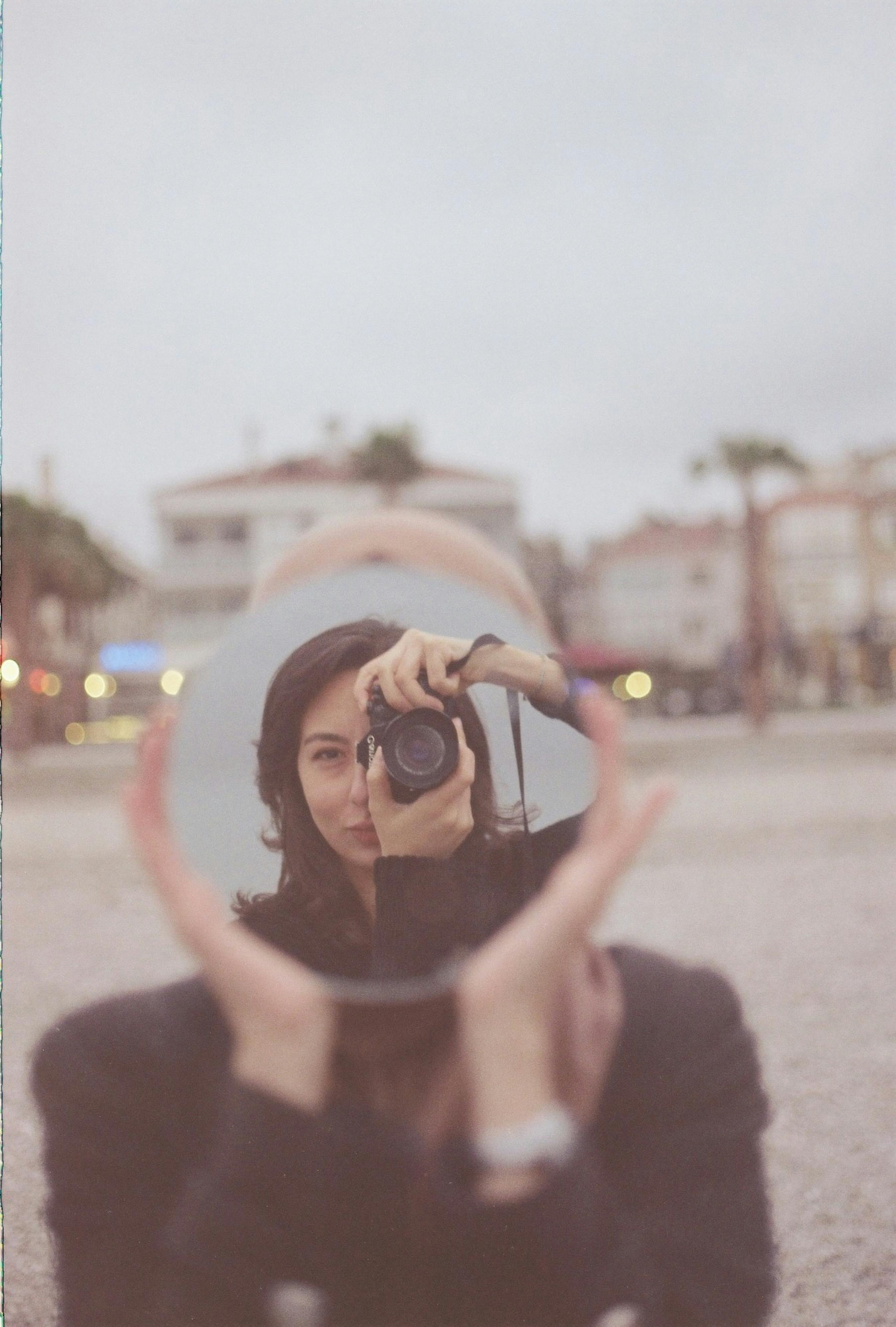 A woman creatively photographs herself using a round mirror in an urban environment.