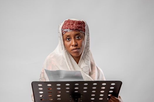 A woman in traditional clothing reads from paper on a white background.