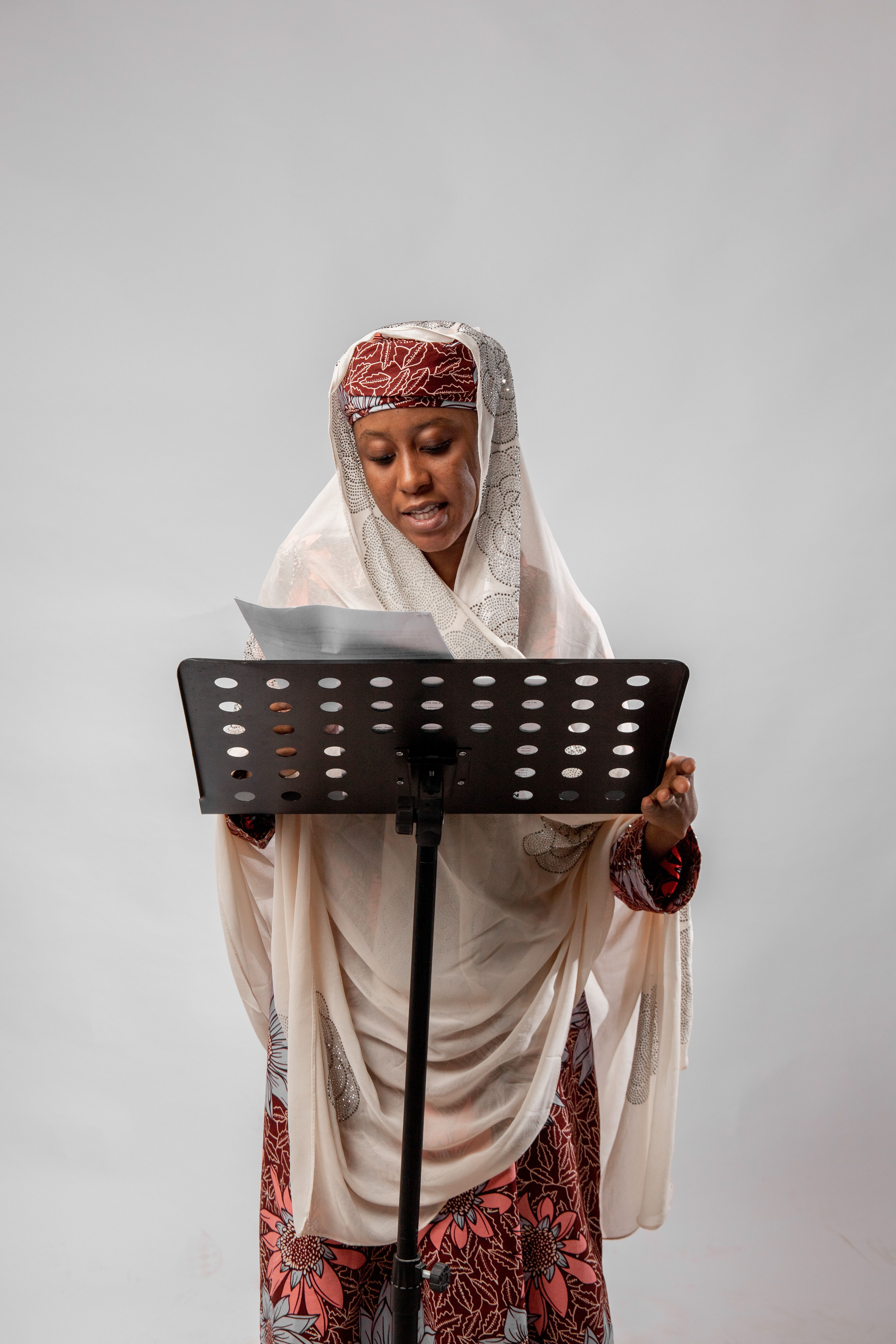 African woman in traditional attire singing, holding sheet music in a studio.