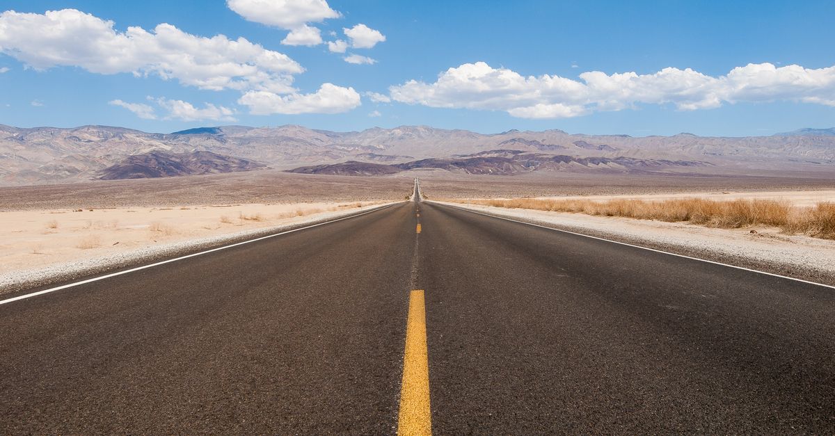 A straight road stretches through the arid desert, framed by mountains under a clear blue sky.