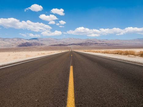A straight road stretches through the arid desert, framed by mountains under a clear blue sky.