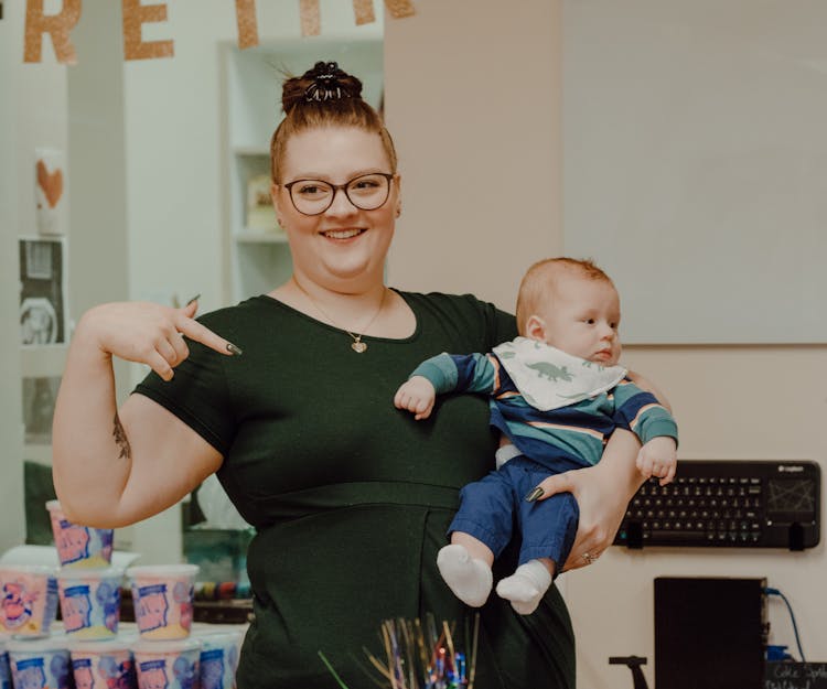 Smiling Woman In Glasses Pointing At Baby On Arms