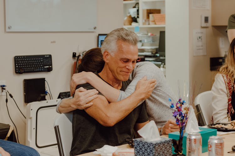 People Hugging Sitting At Table At Celebration In Office