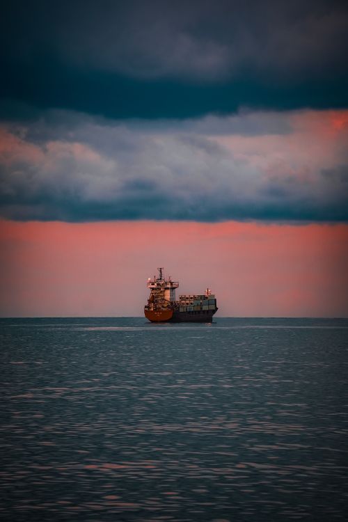 Free A solitary cargo ship sails under a dramatic sky with sunset hues over the calm sea. Stock Photo