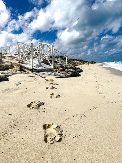 Free Serene beach with footprints leading to a wooden bridge under a bright blue sky. Stock Photo