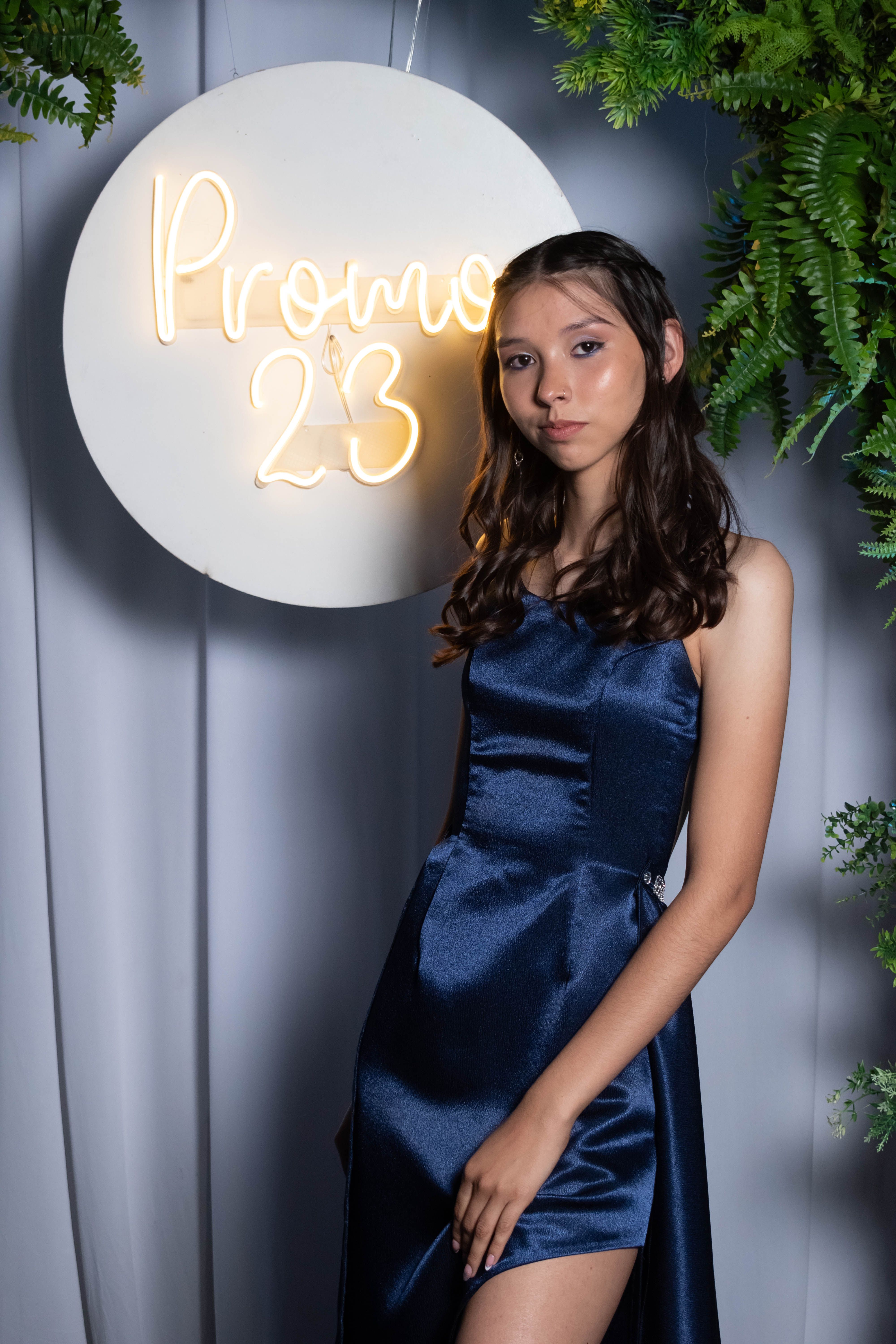 A young woman in an elegant blue dress poses next to a neon 'Prom 23' sign, exuding sophistication.
