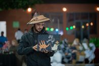 A young man wearing a traditional hat uses a laptop in an outdoor setting during a vibrant night.
