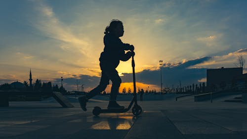 Free Silhouette of a Child Riding on a Scooter in City in the Evening Stock Photo