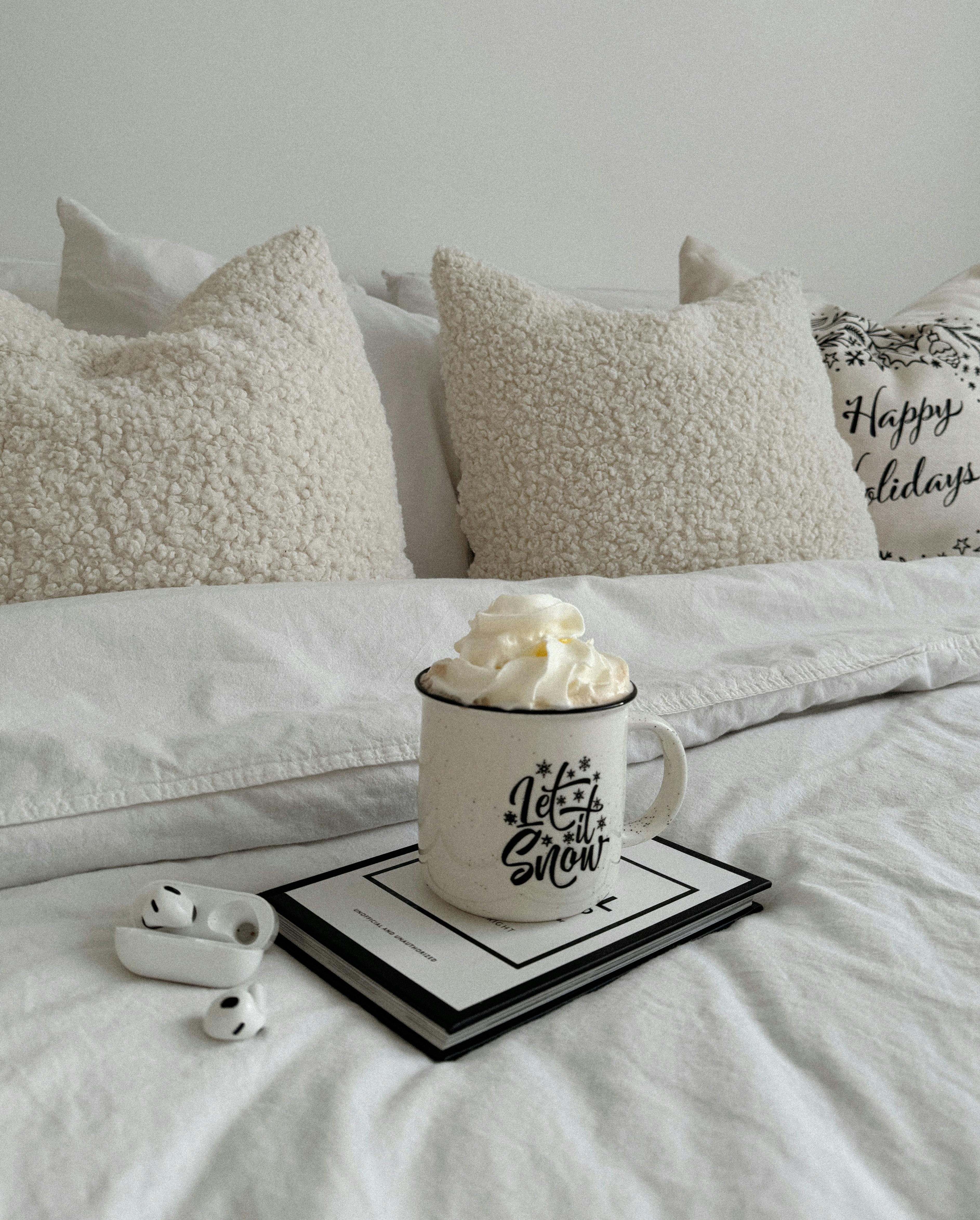 A cozy winter setting with a mug of whipped cream on a bed with pillows and a book.