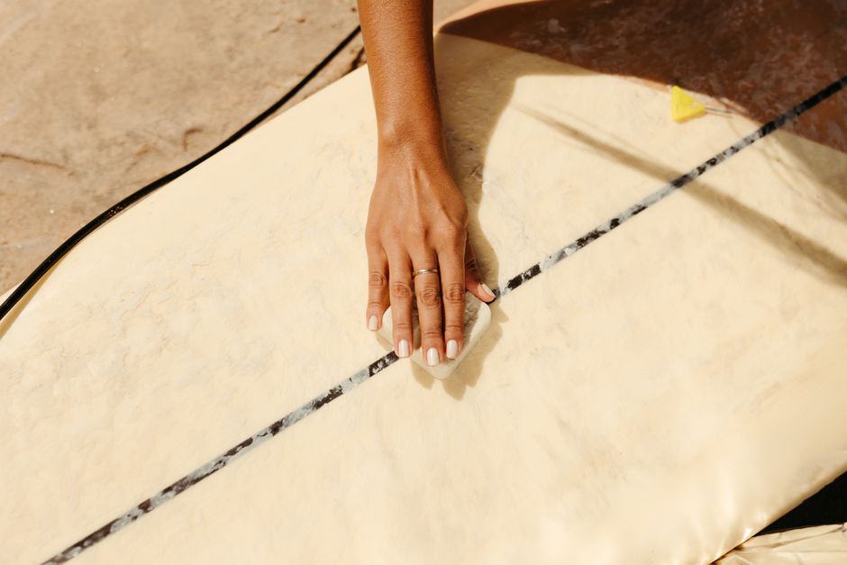 Close-up of a woman applying wax on a surfboard on a sandy beach during summer.