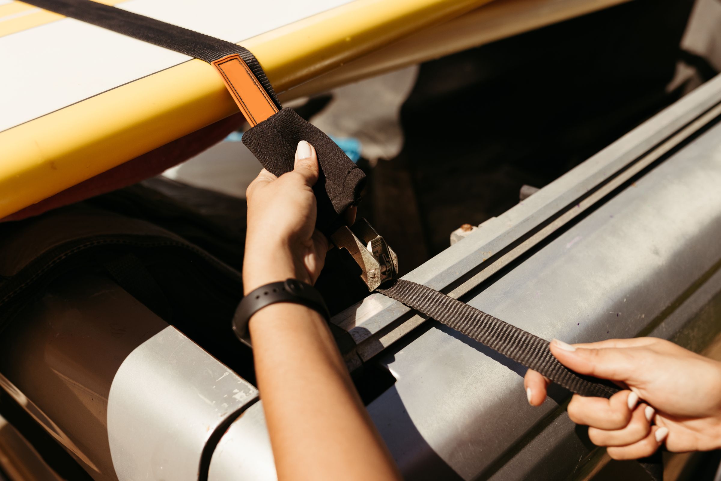 Hands tightening a cam-buckle tie-down strap over a paddleboard on a vehicle roof rack