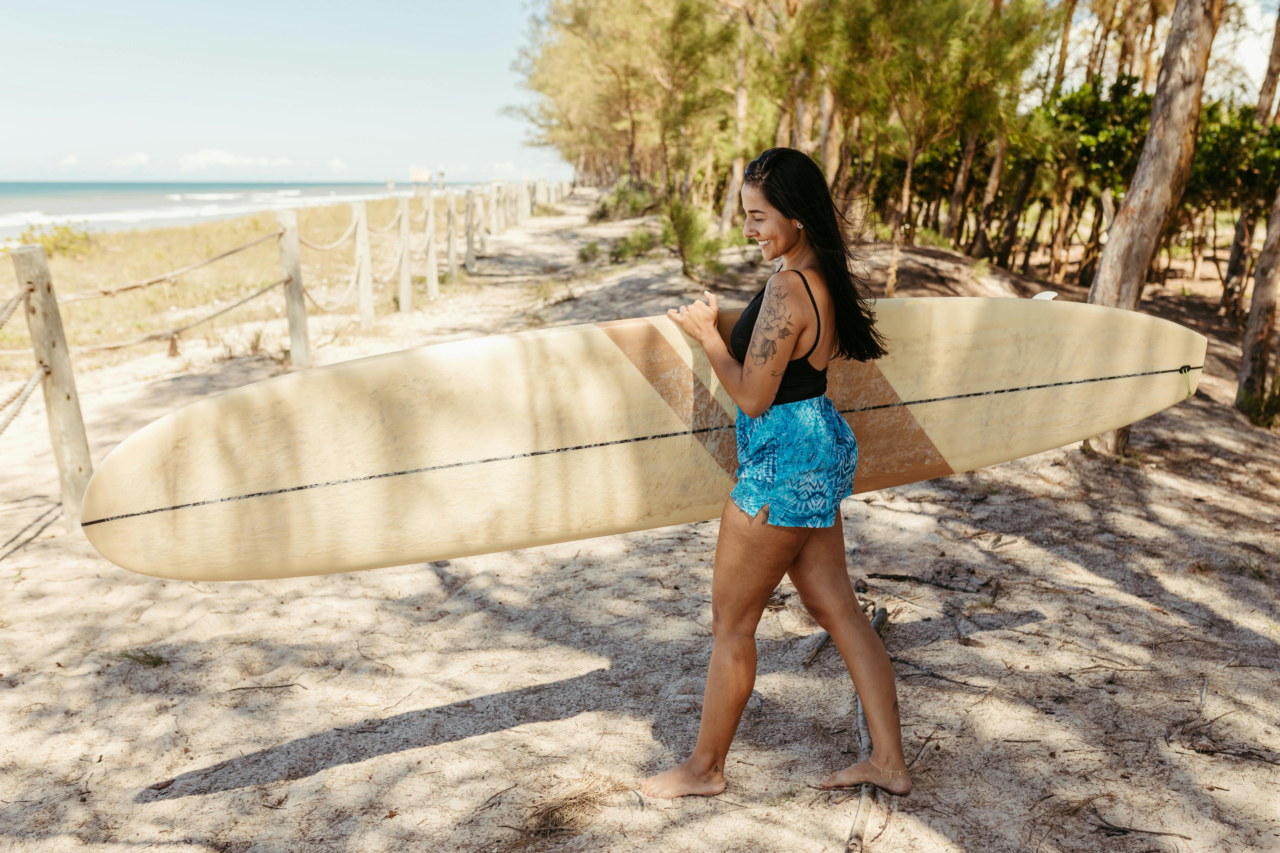Free A woman carries a surfboard along a sunny beach path, exuding relaxation and summer vibes. Stock Photo