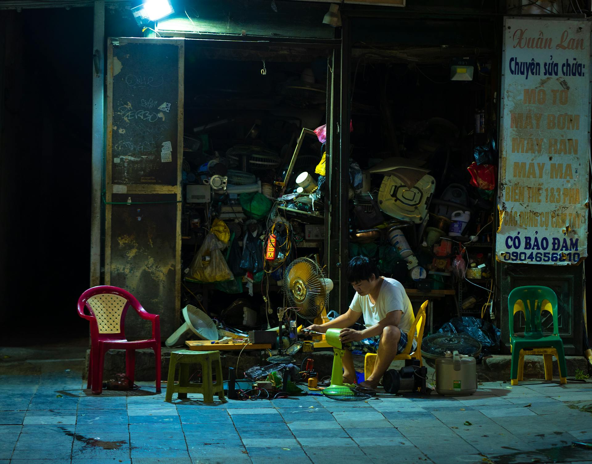 A man repairing household items outside a cluttered shop under streetlights at night.