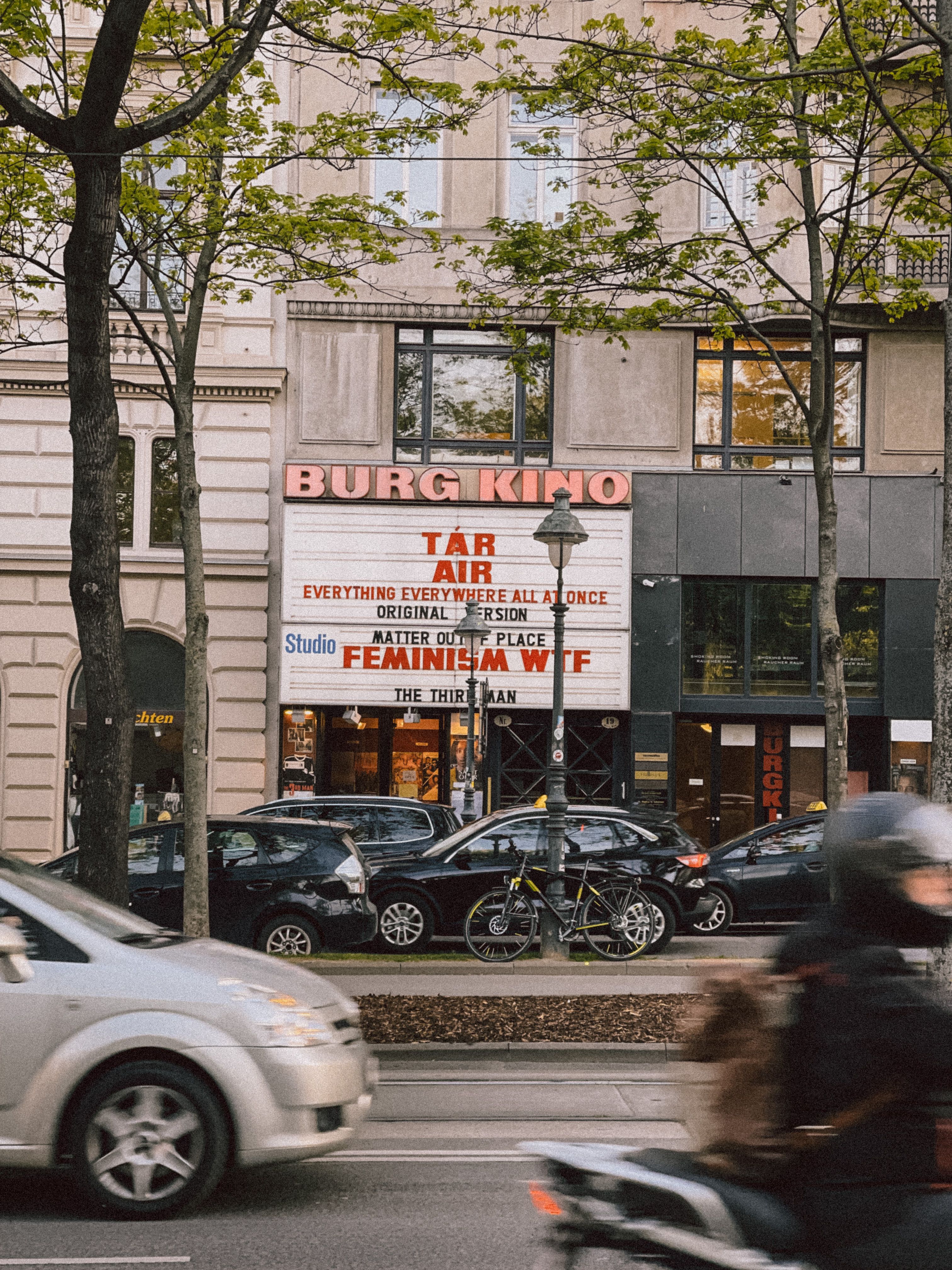 Urban scene in Vienna featuring Burg Kino cinema with trees and vehicles in focus.