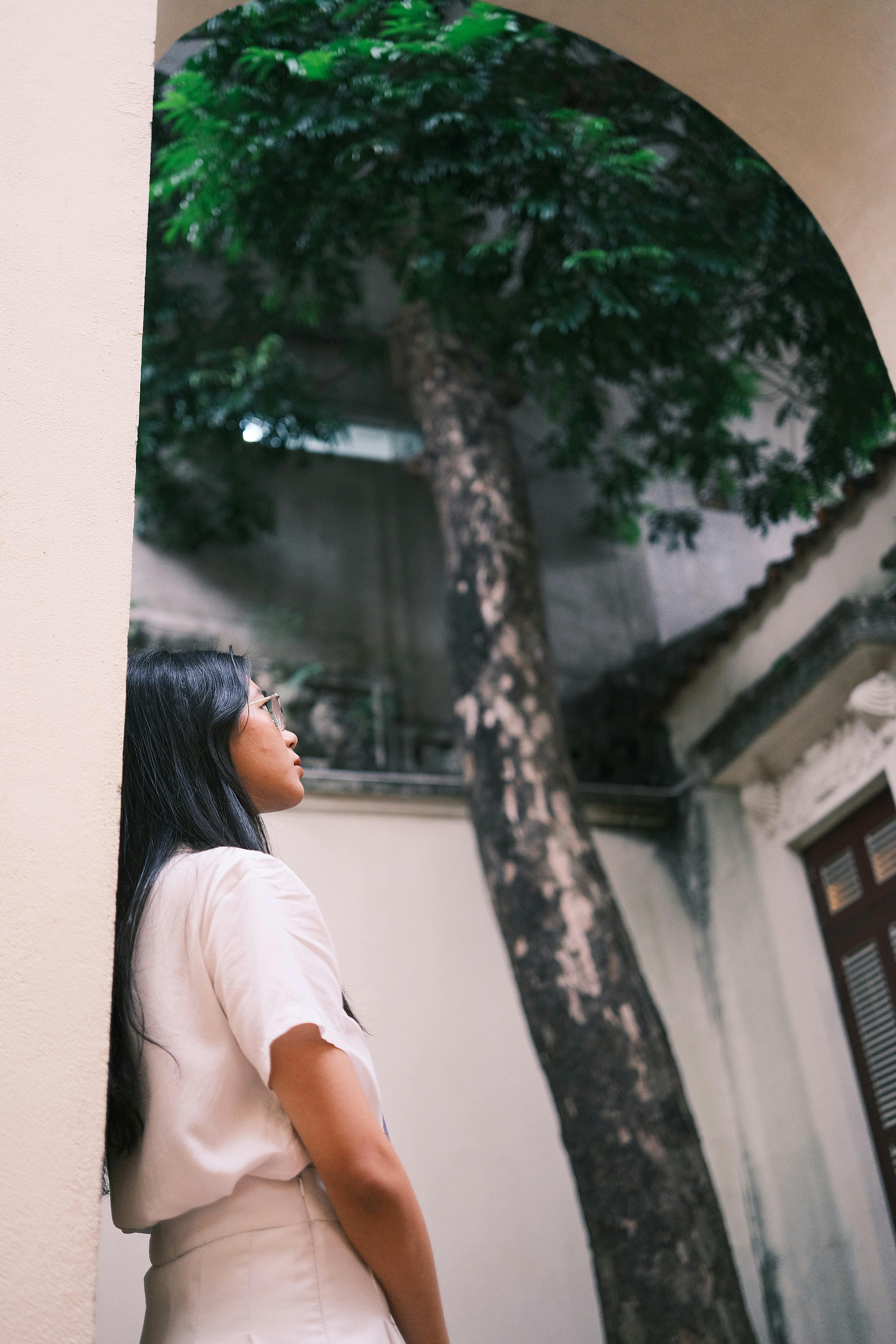 Woman with long hair stands under tree in an urban courtyard, looking upward.