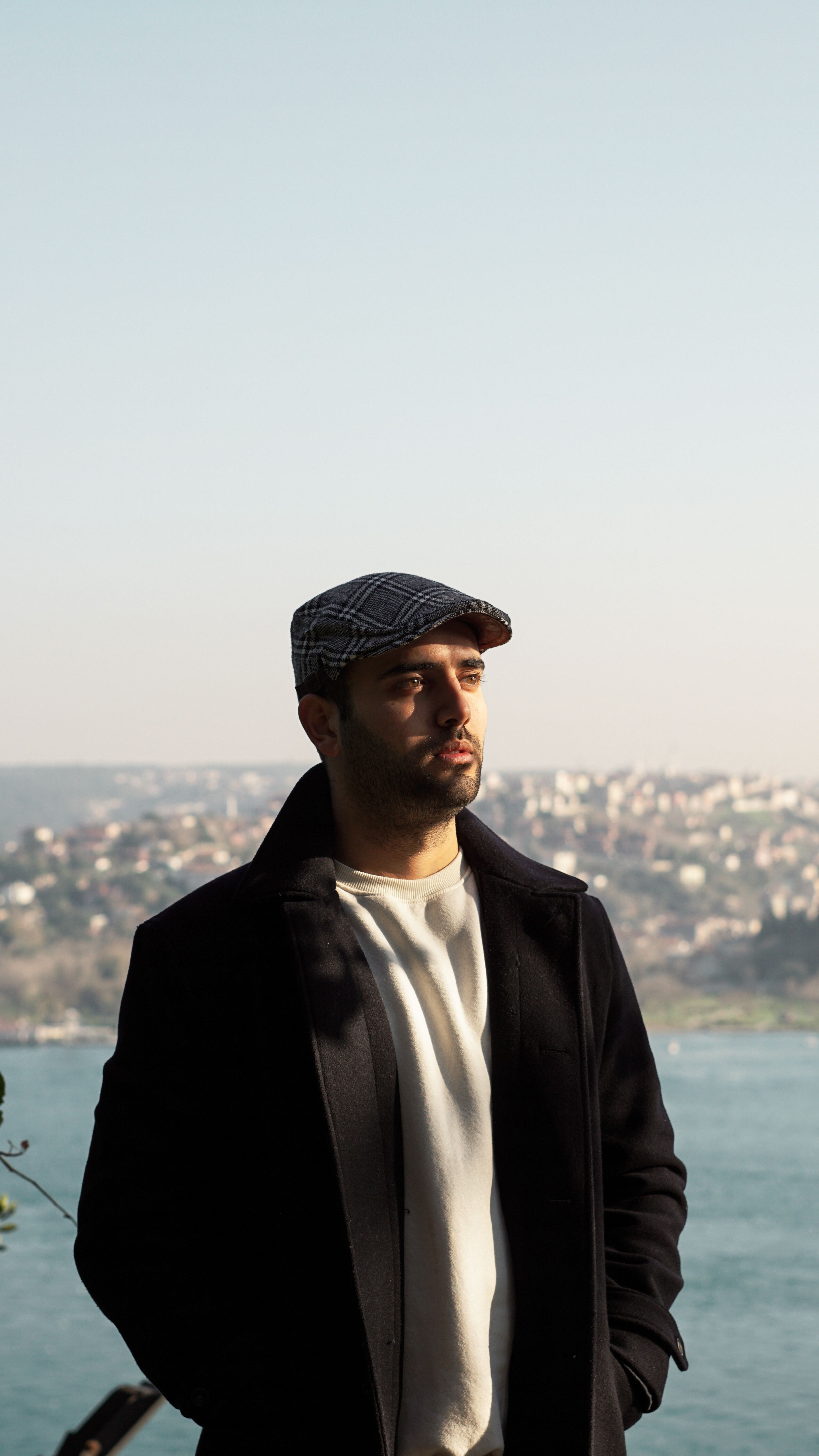 A man wearing a coat and peaked cap stands against the İstanbul skyline with the Bosphorus view.