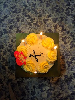 Top view of a birthday cake with yellow rosettes, lit candles, and festive decoration.