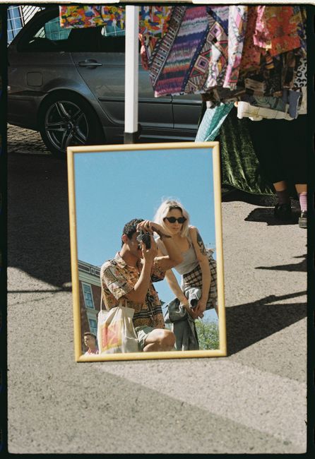 A couple taking photos reflected in a mirror at an outdoor Lisbon market showcasing vibrant summer fashion.