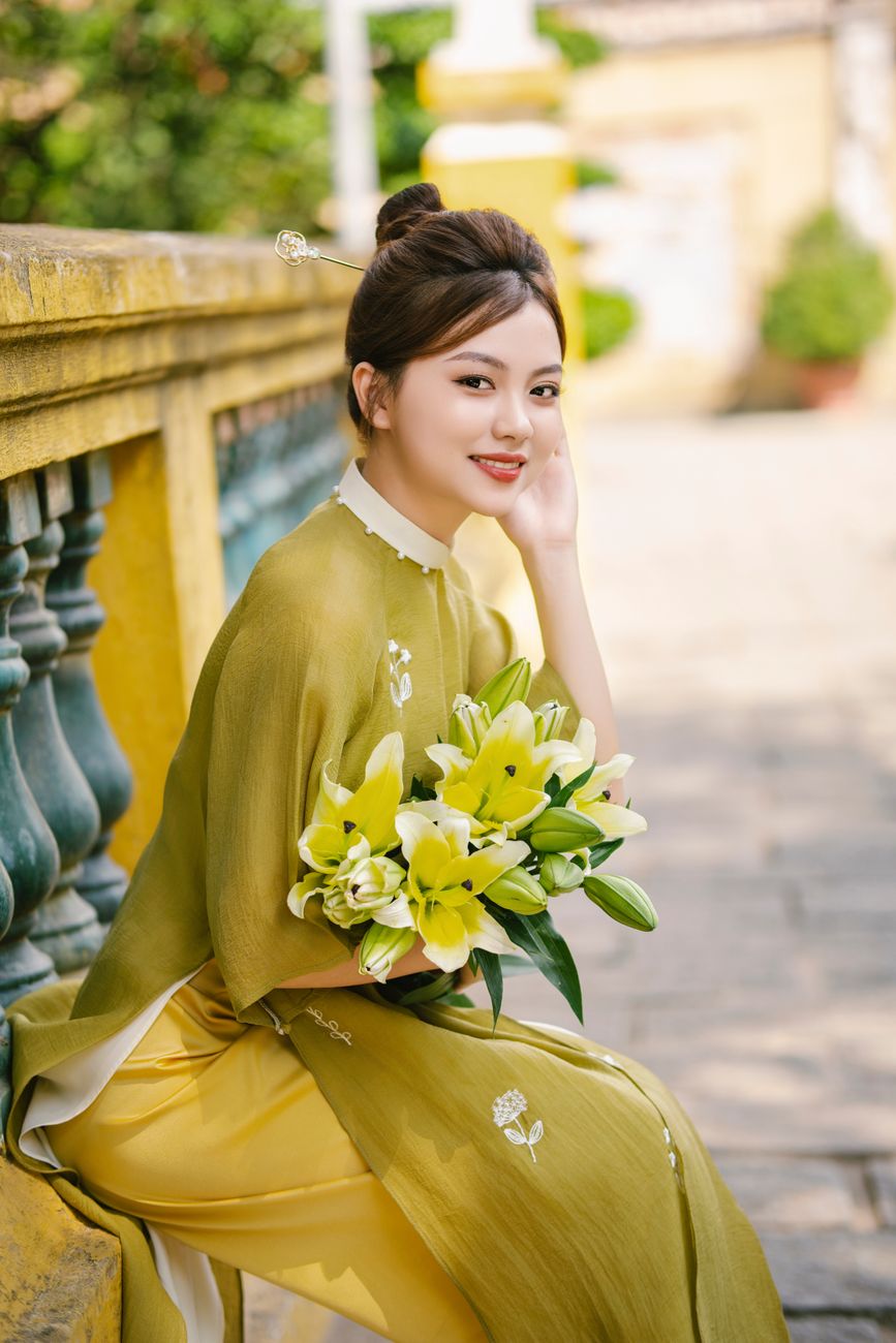 A young Vietnamese woman wearing an ao dai holds a bouquet of lilies, exuding elegance on a sunny day.