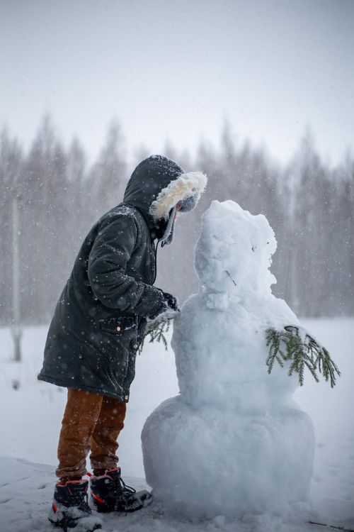 Free A child in a warm coat joyfully builds a snowman during a snowfall in winter. Stock Photo
