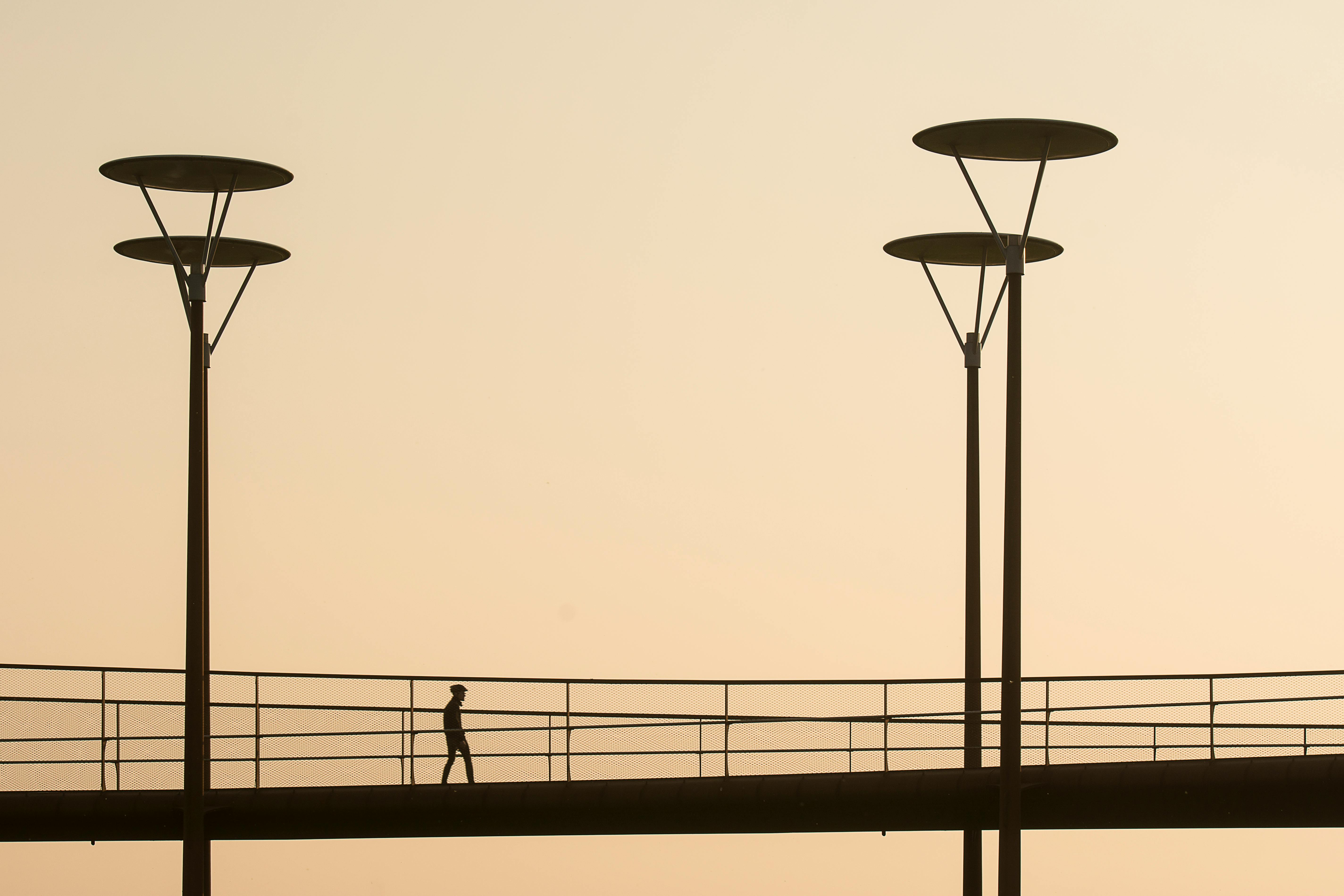A man walks on a silhouetted urban footbridge at sunset with street lamps.