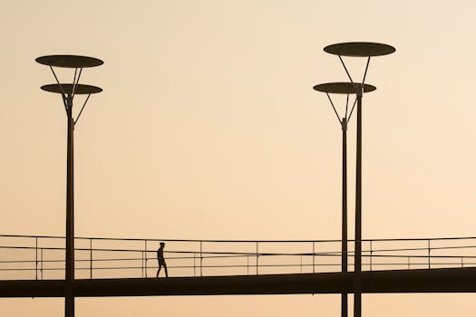 A man walks on a silhouetted urban footbridge at sunset with street lamps.