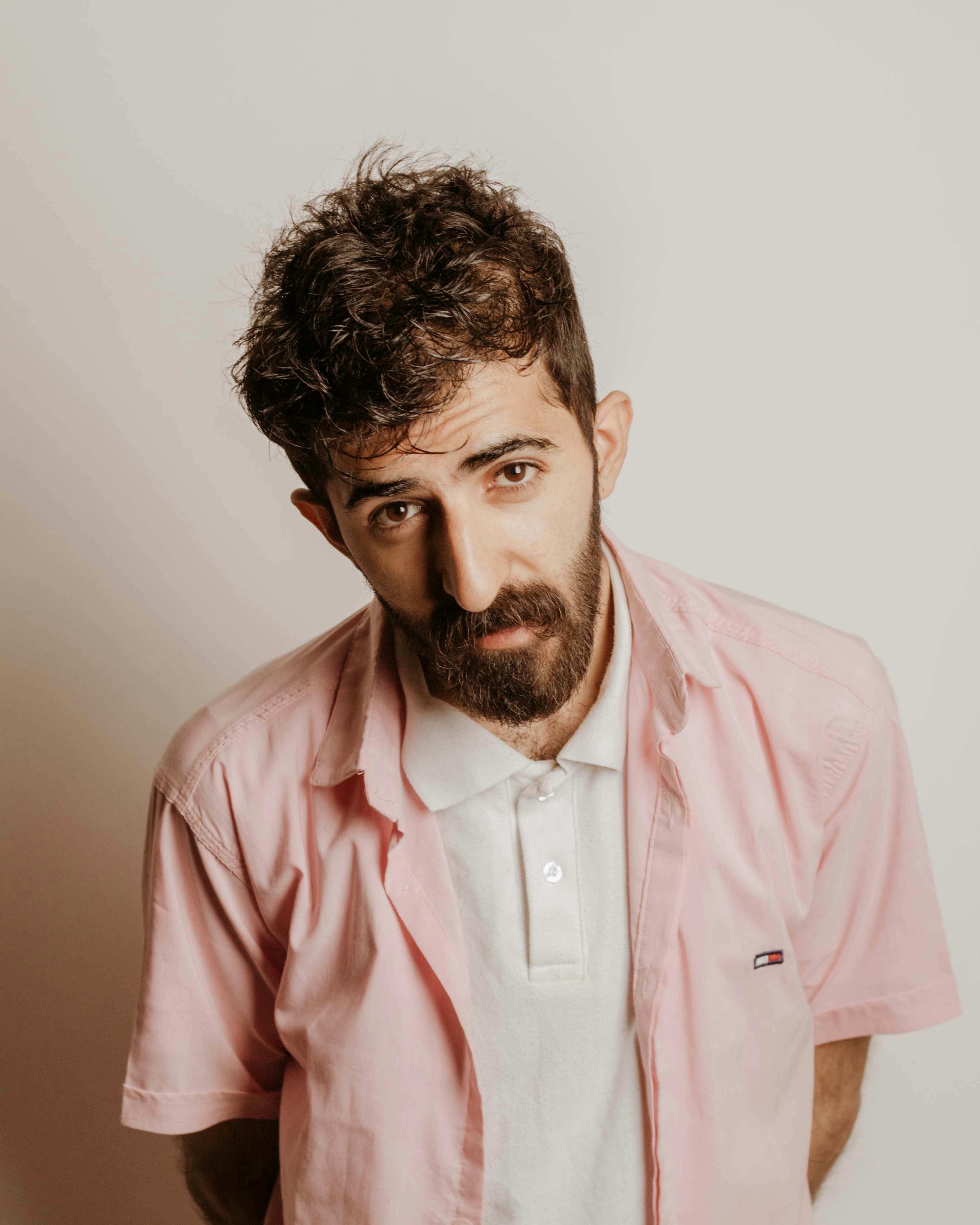 Man with beard wearing a casual pink shirt and white polo posing indoors.