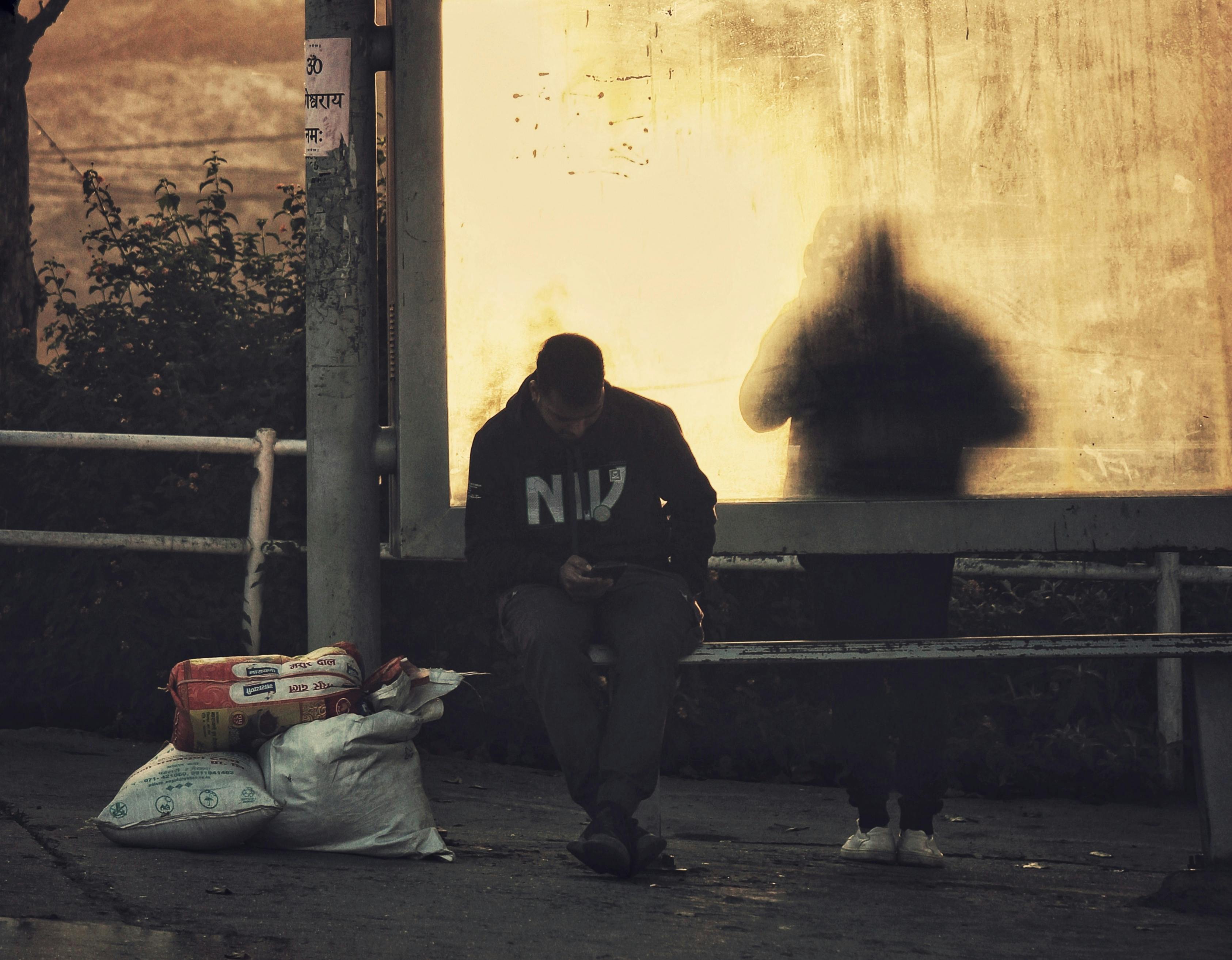 A man in a hoodie uses a smartphone while sitting at a bus stop in Kathmandu, Nepal.