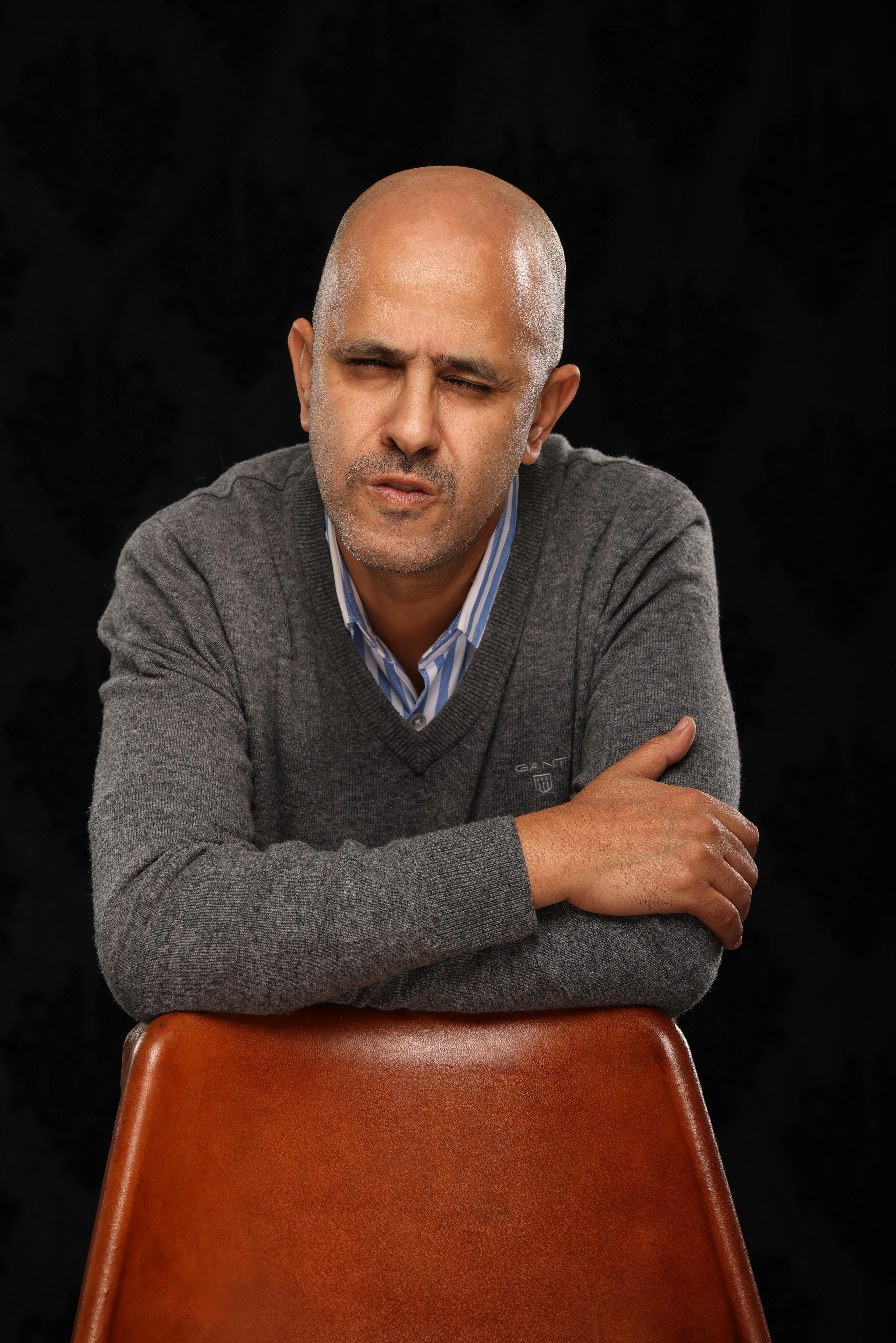 Bald man in a gray sweater leaning on a brown chair against a dark background.