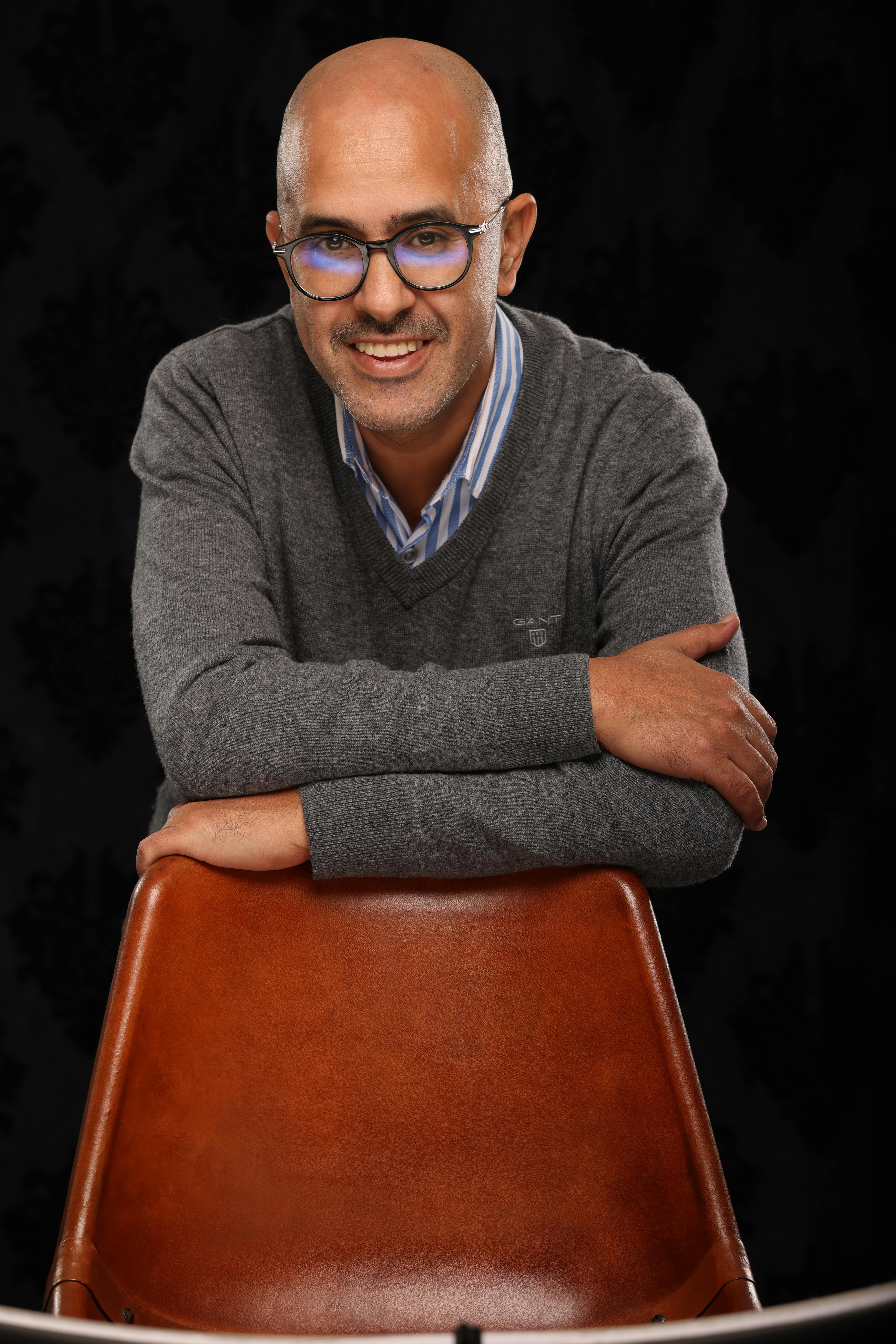 Man with eyeglasses and gray sweater smiling in a studio portrait.
