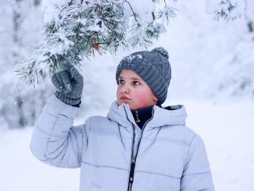 Boy in Beanie and Jacket Touching Snow on Conifer