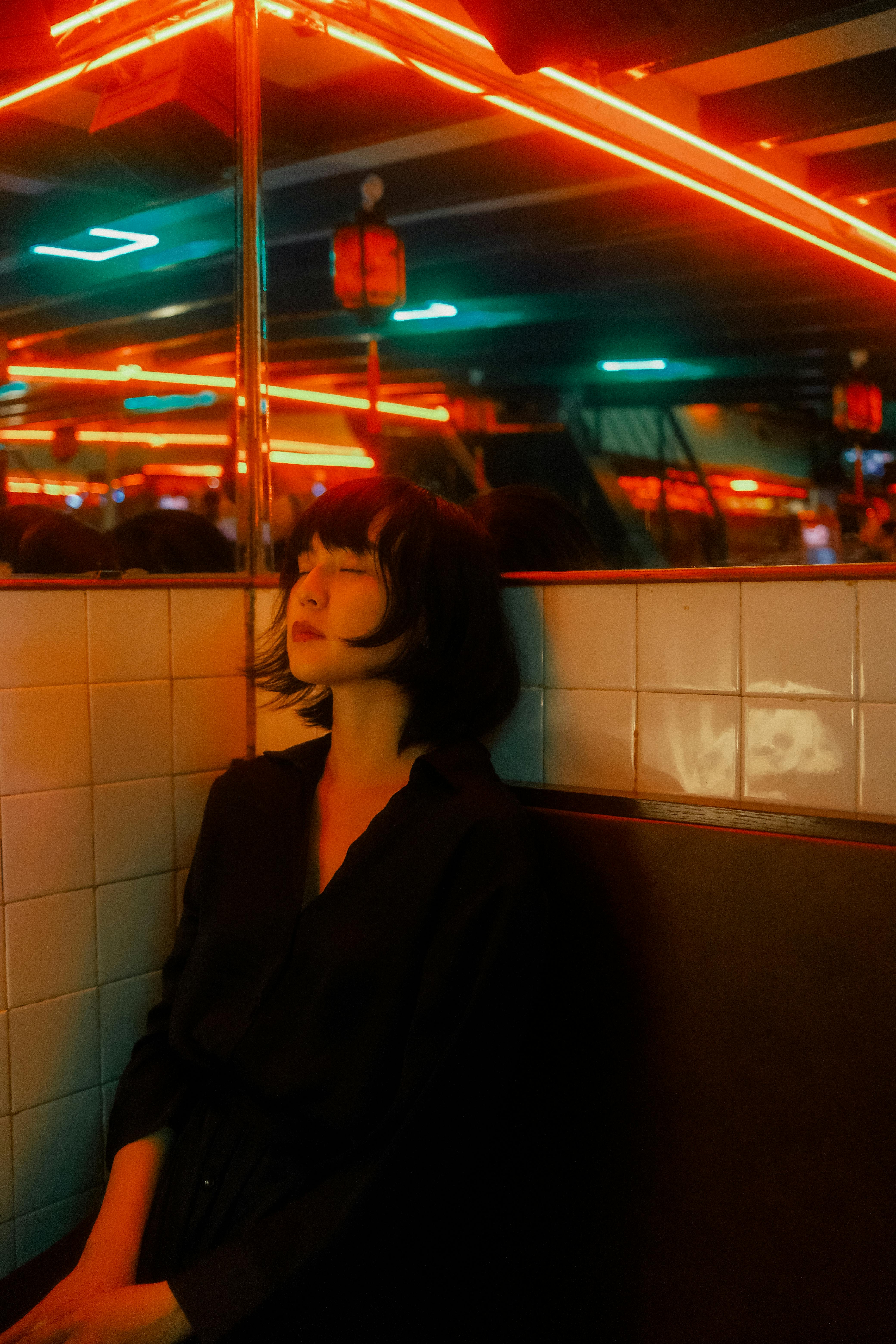 Young woman with closed eyes sitting under vibrant neon lights, creating a moody ambiance.