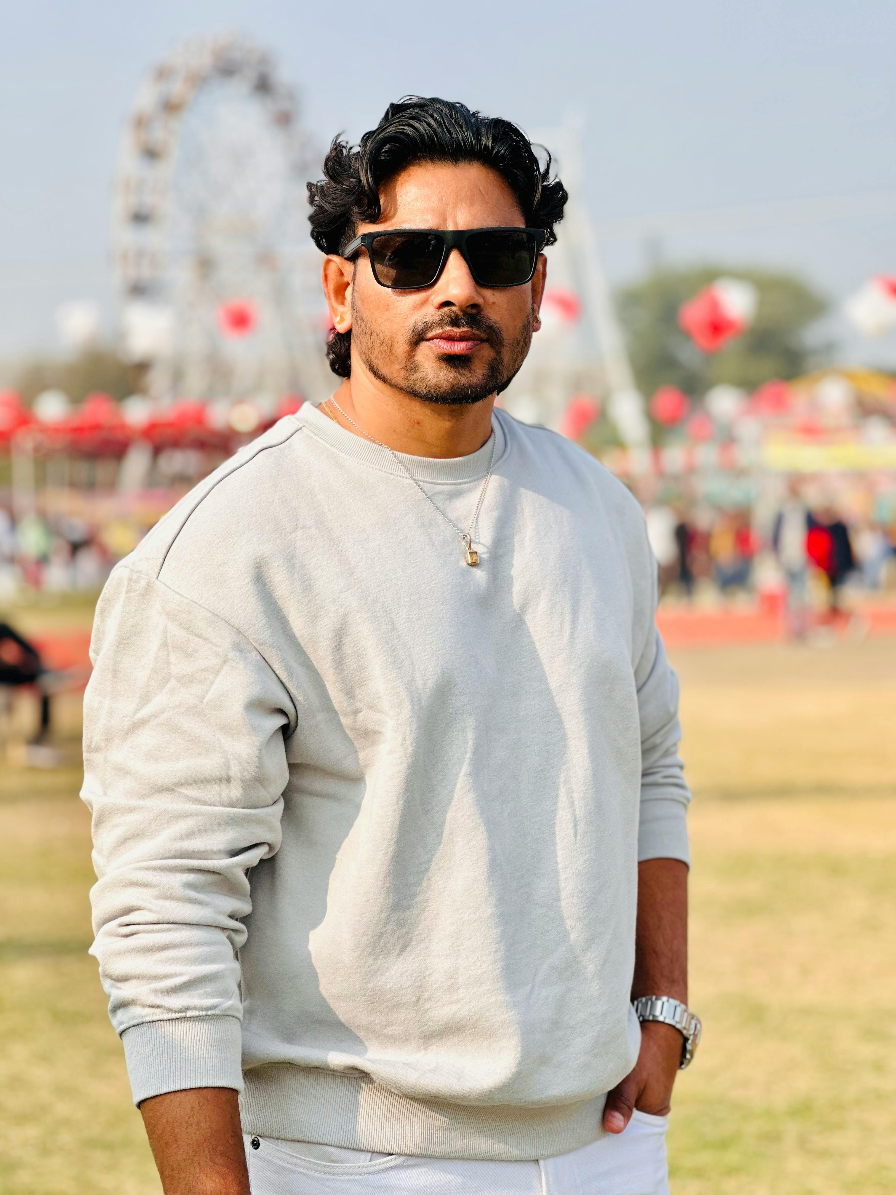 A fashionable man wearing sunglasses and a sweatshirt standing at an outdoor fairground.