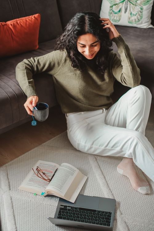 Woman sitting on a rug in the living room, enjoying a cup of tea next to a laptop and an open book.
