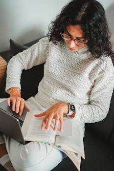 Woman in white sweater sitting on a couch, multitasking with a laptop and book, creating a cozy study environment.