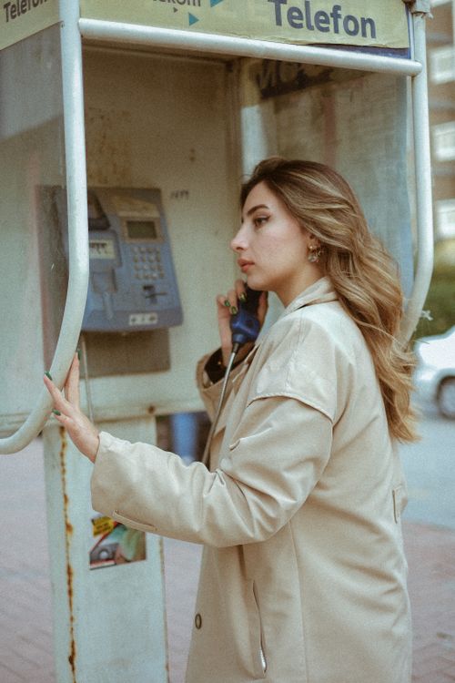 Free Fashionable woman in a trench coat making a call from a vintage public phone booth outdoors. Stock Photo