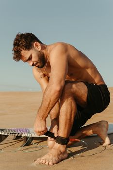 Shirtless man securing surfboard leash to ankle on sandy beach under clear blue sky.