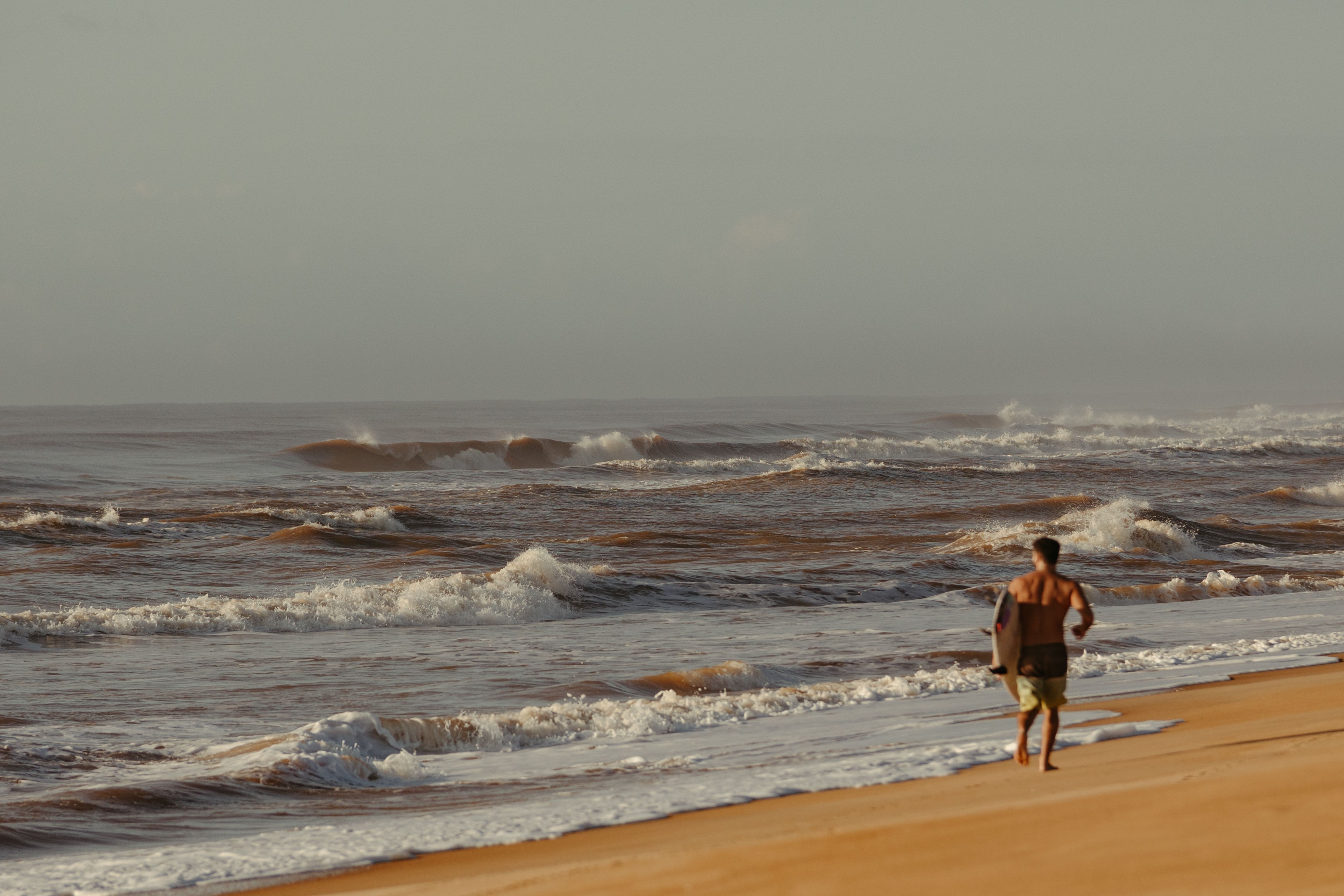 Man strolling on a beach with a surfboard, surrounded by waves under a clear sky.