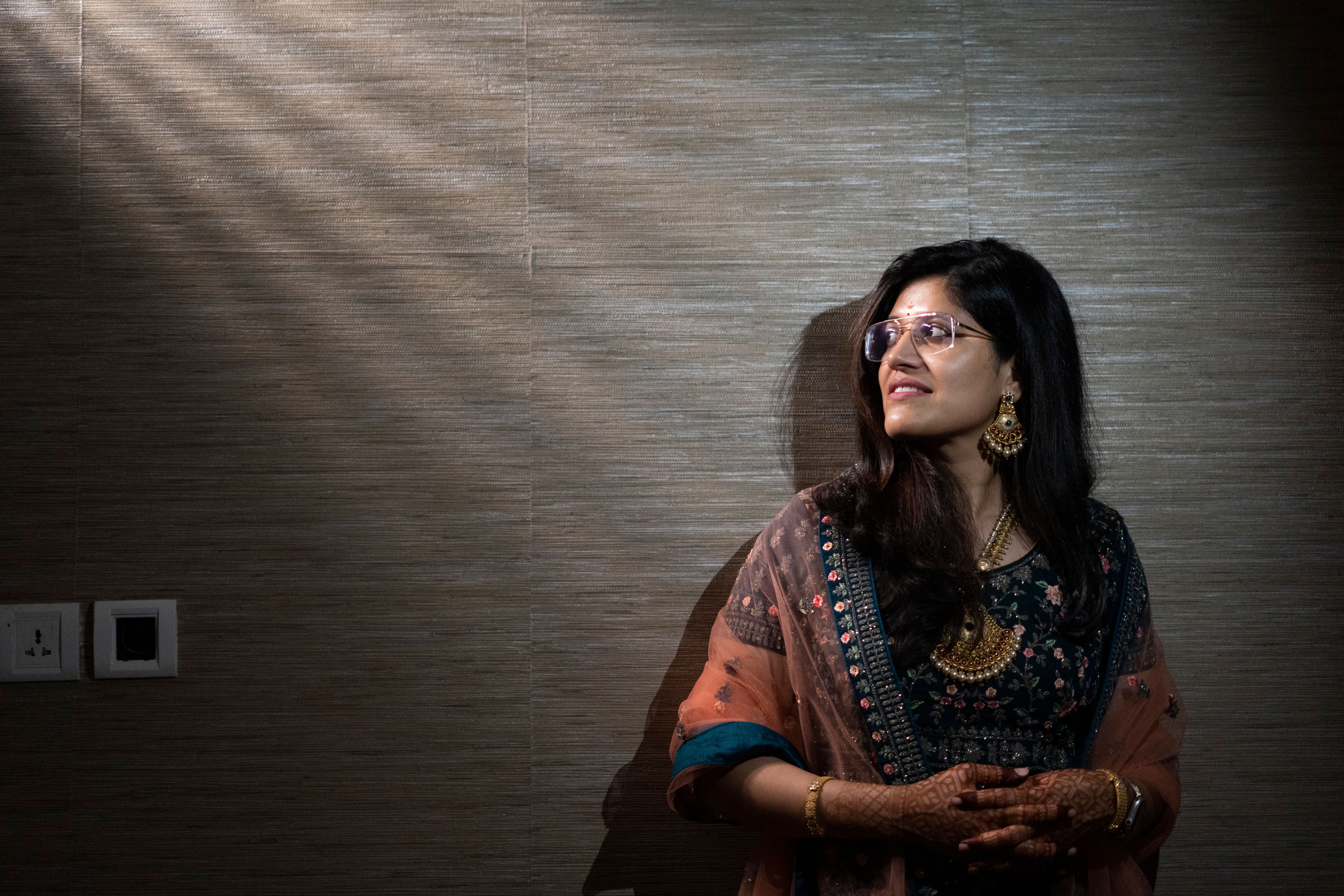 A sunlit portrait of a woman in a traditional saree, wearing eyeglasses, standing indoors.