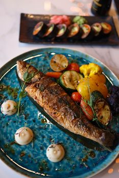 A gourmet meal featuring a grilled fish with assorted roasted vegetables on a blue plate.