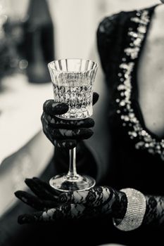 A woman dressed in luxury, wearing lace gloves, holds a crystal glass in a black and white setting.