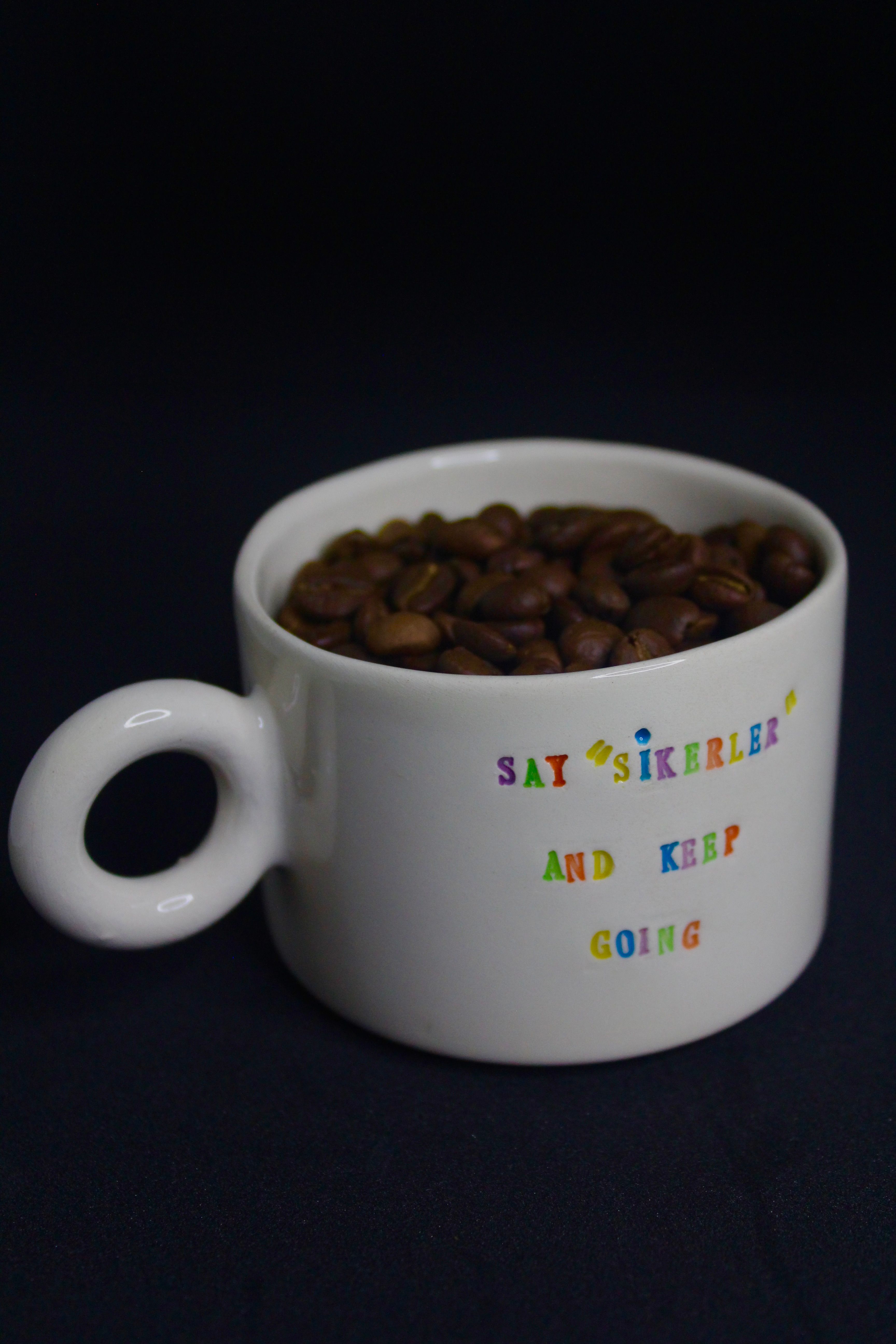 A white mug with colorful text filled with coffee beans against a black background.