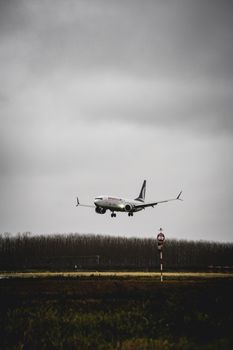 A commercial airplane descends onto a runway against a backdrop of cloudy, overcast skies and distant tree line.