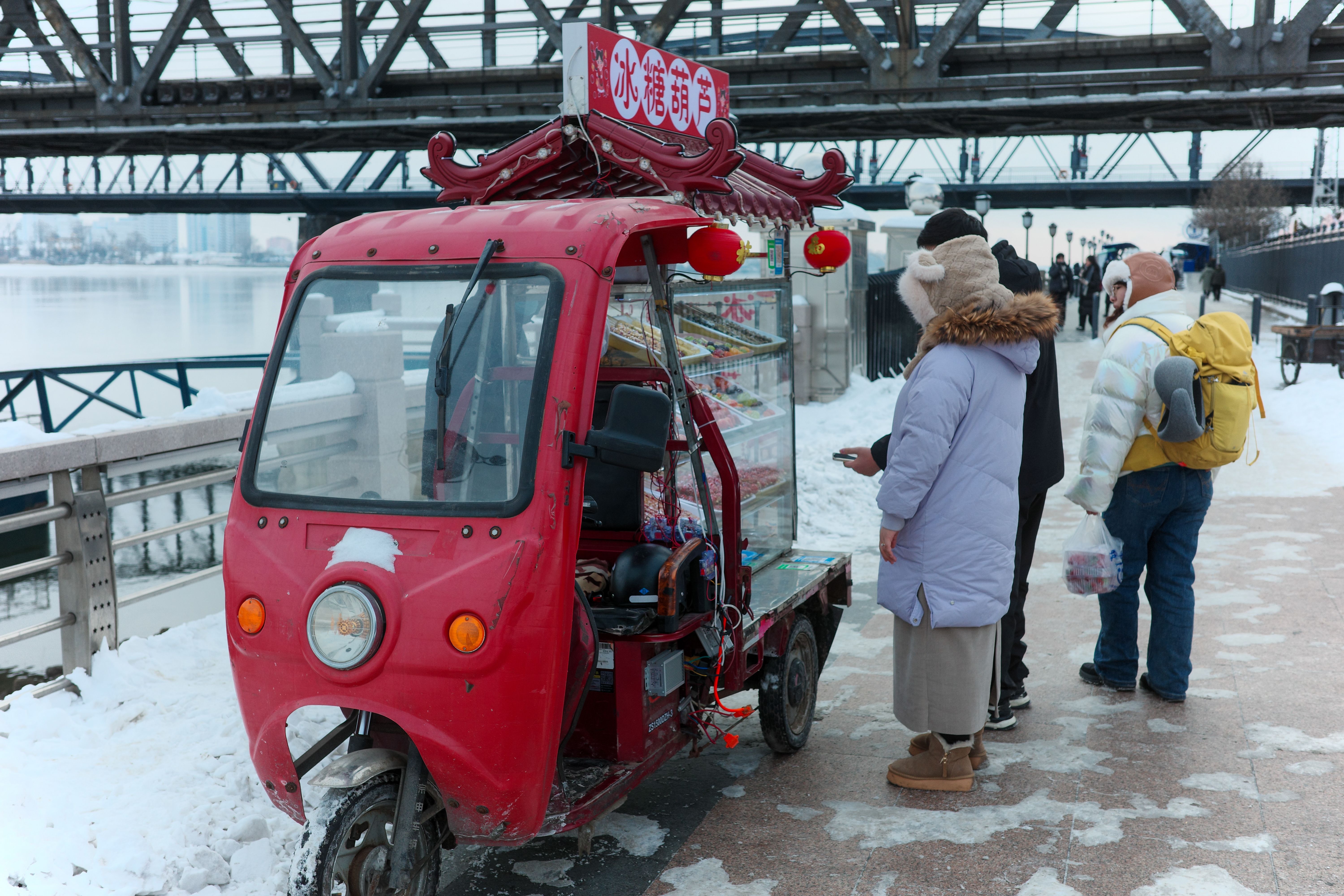 Free People buy from a street vendor near a snowy river with a red cart. Winter scene with bridge backdrop. Stock Photo