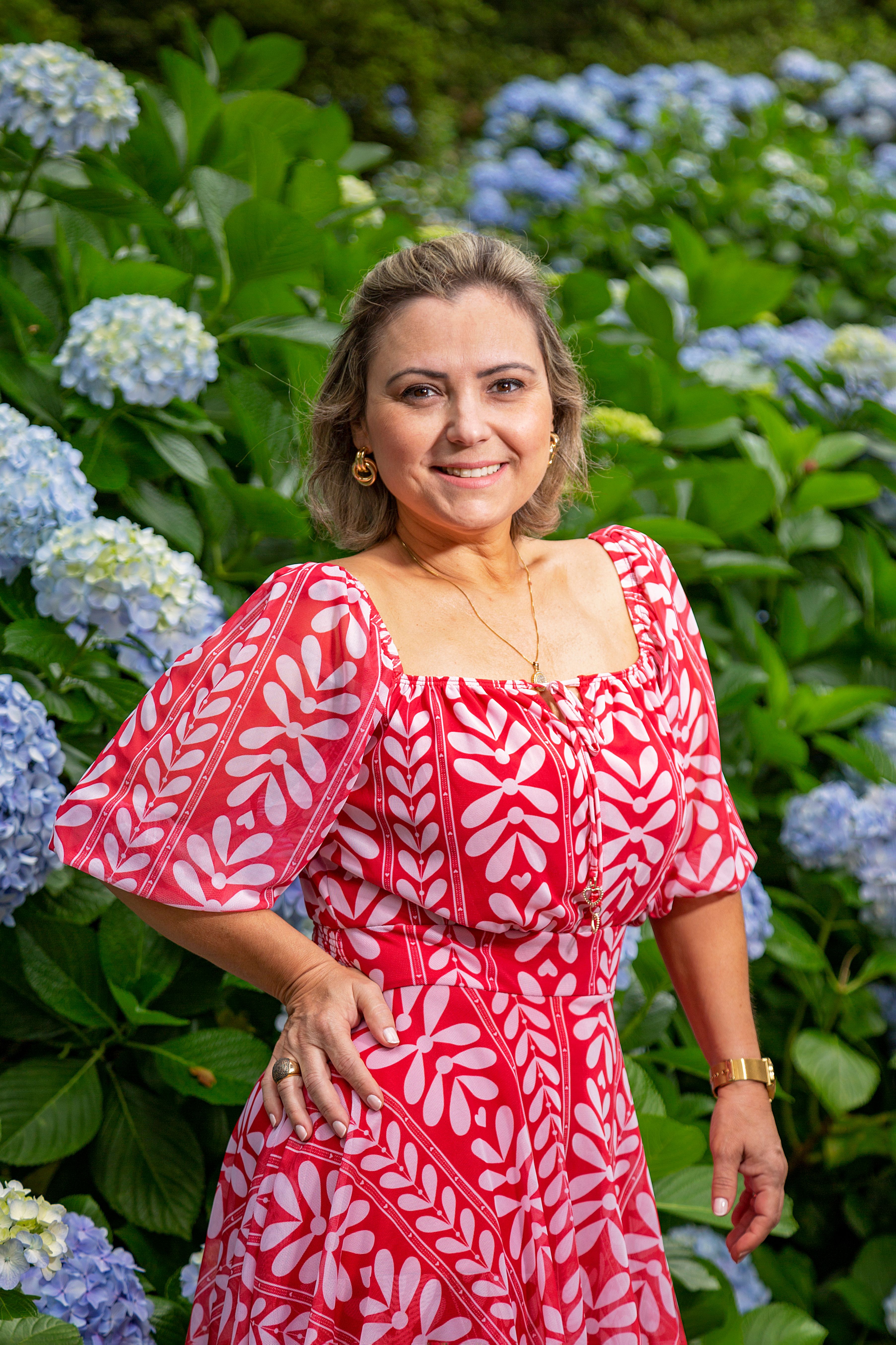 A smiling woman in a red floral dress poses outdoors with vibrant hydrangeas.